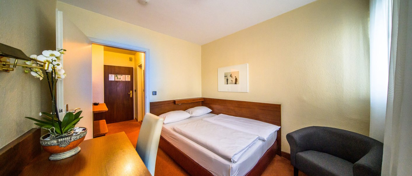 Ein einfaches Hotelzimmer mit Doppelbett, Schreibtisch, Sessel und Bild an der Wand. Eine Orchidee steht auf dem Tisch., © PLAZA INN Rieker Stuttgart Hauptbahnhof Ein einfaches Hotelzimmer mit Doppelbett, Schreibtisch, Sessel und Bild an der Wand. Eine Orchidee steht auf dem Tisch., © PLAZA INN Rieker Stuttgart Hauptbahnhof