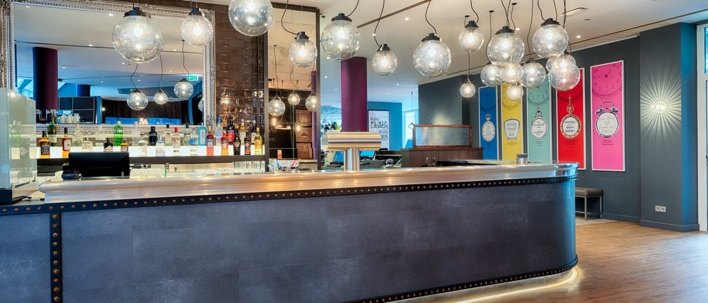 Modern bar with hanging glass ball lamps, mirror and colorful wall decorations. Bottles and glasses are placed on the bar., © Premier Inn Germany Modern bar with hanging glass ball lamps, mirror and colorful wall decorations. Bottles and glasses are placed on the bar., © Premier Inn Germany