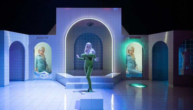 A person in a green outfit stands on a futuristic stage with illuminated arches and pictures in the background., © Württembergische Staatstheater Stuttgart A person in a green outfit stands on a futuristic stage with illuminated arches and pictures in the background., © Württembergische Staatstheater Stuttgart