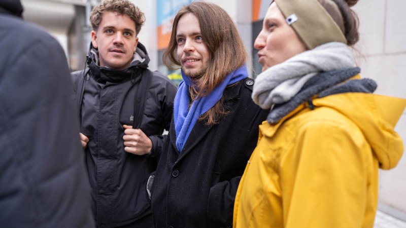 Drei Personen in Winterkleidung unterhalten sich draußen. Sie tragen Jacken und Schals, während sie in einer städtischen Umgebung stehen., © Elisa Mauruschat Drei Personen in Winterkleidung unterhalten sich draußen. Sie tragen Jacken und Schals, während sie in einer städtischen Umgebung stehen., © Elisa Mauruschat