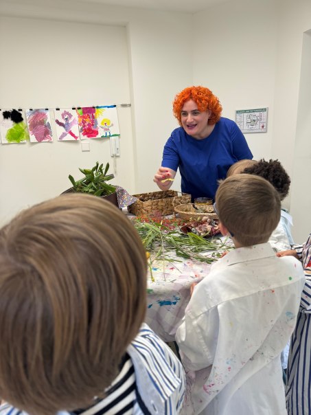 Eine Kursleiterin mit orangefarbenem Haar leitet einen Kunstkurs f&uuml;r Kinder. Auf dem Tisch liegen Pflanzen und Kunstmaterialien. Kinder in Malkitteln h&ouml;ren zu., &copy; Adolf H&ouml;lzel Stiftung