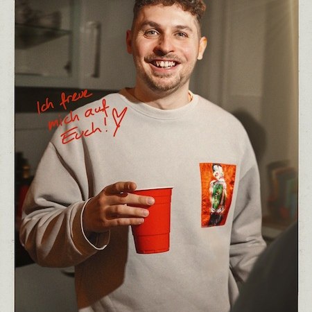 Mann mit lockigem Haar h&auml;lt roten Becher, l&auml;chelt. Text: 'Einer dieser Abende mit Fabian Lampert'. Hintergrund unscharf, gem&uuml;tliche Atmosph&auml;re., &copy; Rosenau Kultur e.V.