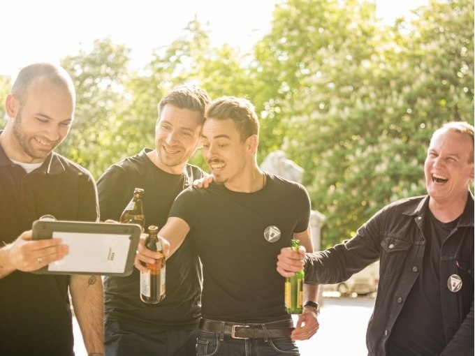 Vier Männer lachen und stoßen mit Bierflaschen an, während sie auf ein Tablet schauen. Im Hintergrund sind Bäume zu sehen., © ExitGames Stuttgart Vier Männer lachen und stoßen mit Bierflaschen an, während sie auf ein Tablet schauen. Im Hintergrund sind Bäume zu sehen., © ExitGames Stuttgart