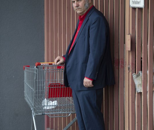 A man in a blue suit with a red shirt stands seriously next to a shopping cart in front of a wooden gate., &copy; Rosenau Kultur e.V.