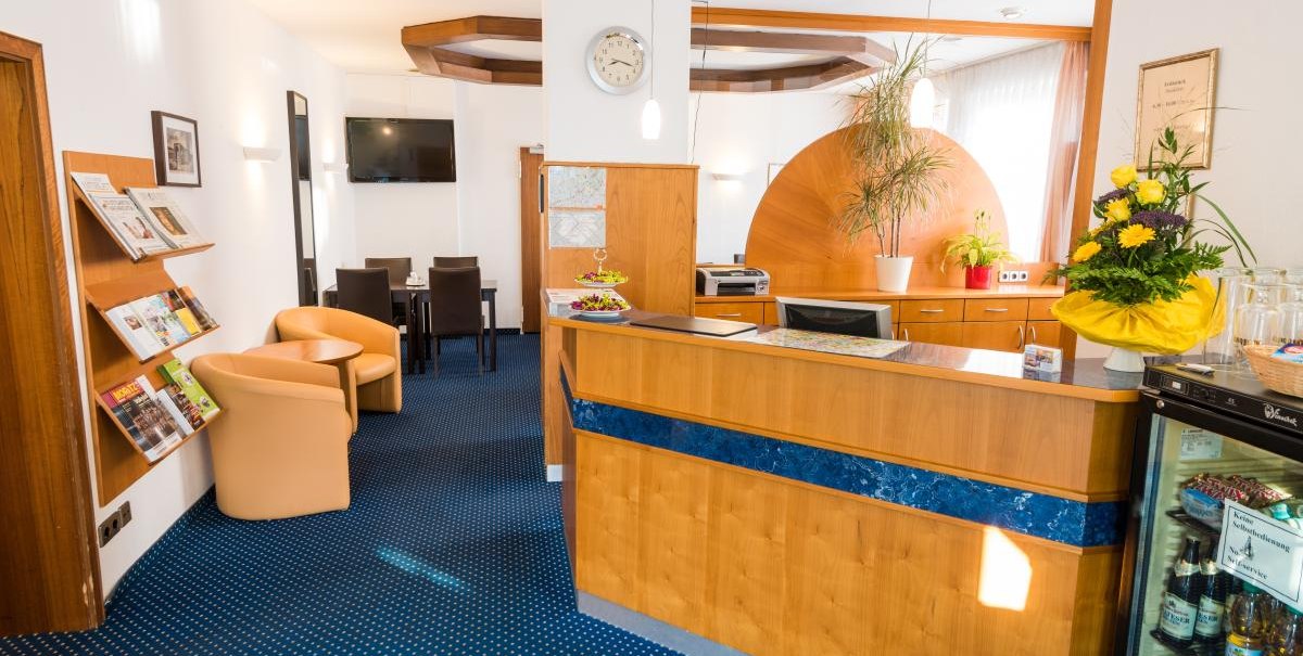 Hotelrezeption mit Holztresen, blauen Teppichboden, Sitzbereich mit Sesseln, Pflanzen und Zeitschriftenständer., © ARNOTEL GmbH