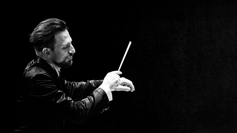 Ein Dirigent führt mit einem Taktstock eine Probe durch. Der Hintergrund ist dunkel, was den Fokus auf seine konzentrierte Gestik lenkt., © Württembergische Staatstheater Stuttgart