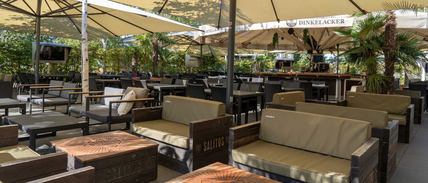 Gemütlicher Außenbereich mit Sofas, Tischen, Sonnenschirmen und Palmen. Ein Fernseher und eine Bar sind ebenfalls sichtbar., © Palm Beach Stuttgart GmbH