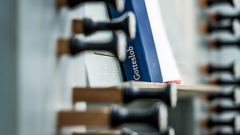 Ein Gotteslob-Buch steckt zwischen den Registern einer Orgel. Die Aufnahme fokussiert auf die Registerzüge und das Buch., © Staatliche Hochschule für Musik und Darstellende Kunst Ein Gotteslob-Buch steckt zwischen den Registern einer Orgel. Die Aufnahme fokussiert auf die Registerzüge und das Buch., © Staatliche Hochschule für Musik und Darstellende Kunst