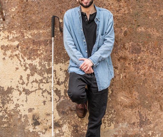 Ein Mann mit Bart und Mütze lehnt lächelnd an einer rustikalen Wand. Er trägt eine Jeansjacke und hält einen Blindenstock., © Rosenau Kultur e.V. Ein Mann mit Bart und Mütze lehnt lächelnd an einer rustikalen Wand. Er trägt eine Jeansjacke und hält einen Blindenstock., © Rosenau Kultur e.V.