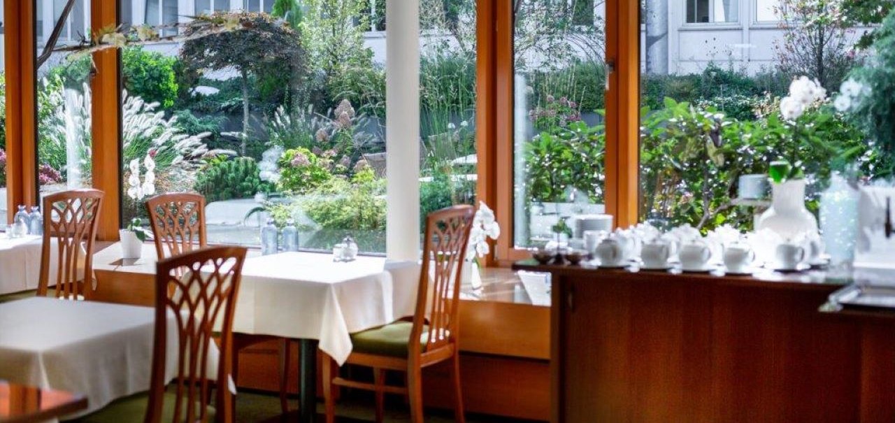 Bright breakfast room with wooden furniture, white tablecloths and a view of a green garden through large windows., © Kronen Hotel GmbH
