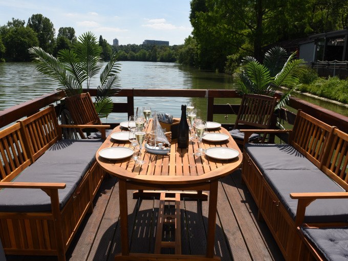 A wooden table on a raft, set with plates and glasses, surrounded by wooden benches and plants, on a river in sunny weather., &copy; Cool-Tours StattReisen