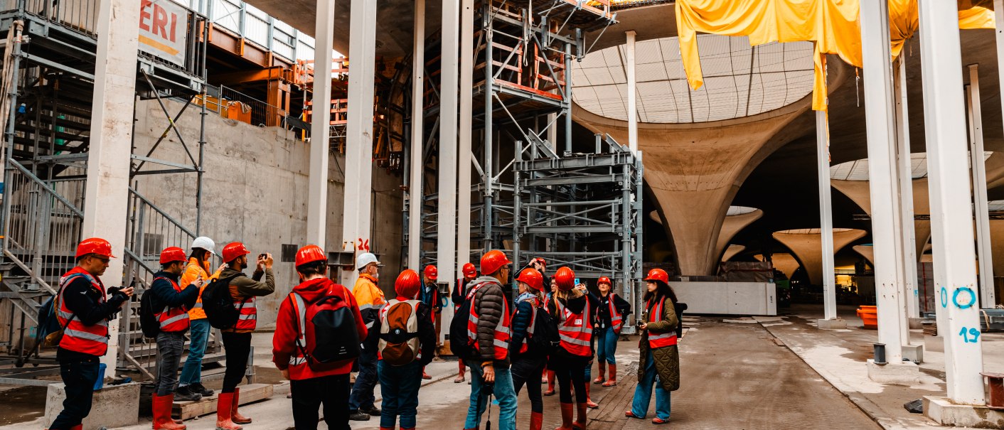 Menschen mit roten Helmen und Stiefeln besichtigen eine Baustelle mit großen Betonpfeilern und Gerüsten. Gelbe Planen hängen von der Decke., © SMG Stuttgart Marketing GmbH - Sarah Schmid