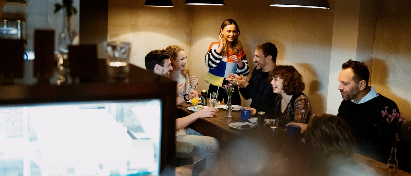 Eine Gruppe von Menschen sitzt in einem Caf&eacute; um einen Tisch und lacht. Eine Frau steht und spricht mit den anderen. Die Atmosph&auml;re ist gem&uuml;tlich., &copy; Stuttgart Marketing GmbH, Alwin Maigler