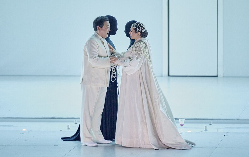 Zwei Personen in weißen Kostümen stehen sich gegenüber, begleitet von zwei schwarz gekleideten Figuren auf einer Bühne., © Württembergische Staatstheater Stuttgart Zwei Personen in weißen Kostümen stehen sich gegenüber, begleitet von zwei schwarz gekleideten Figuren auf einer Bühne., © Württembergische Staatstheater Stuttgart