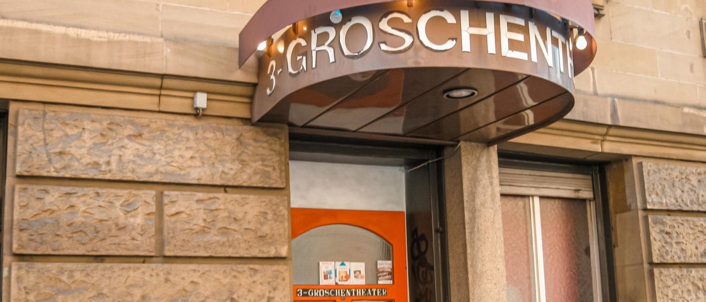 Eingang des 3-Groschen-Theaters mit einem Vordach und Leuchtschrift. Die Fassade ist aus Stein, und es gibt ein kleines Schaufenster mit Plakaten., © Dreigroschentheater