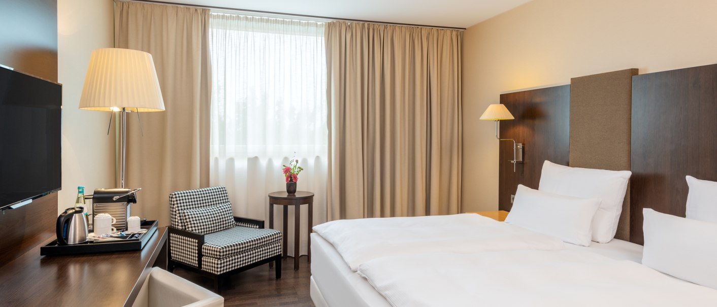 Modernes Doppelzimmer mit gro&szlig;em Bett, Schreibtisch, Sessel und Fenster mit beigen Vorh&auml;ngen. Ein Fernseher und eine Stehlampe sind ebenfalls vorhanden., &copy; NH Hotel Stuttgart Airport