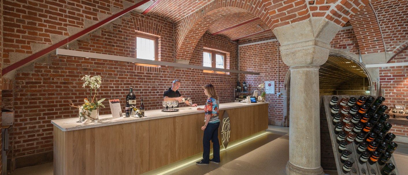 A bar in a wine cellar with brick walls. A man pours wine for a woman. Wine bottles are displayed on the counter., © Collegium Wirtemberg A bar in a wine cellar with brick walls. A man pours wine for a woman. Wine bottles are displayed on the counter., © Collegium Wirtemberg