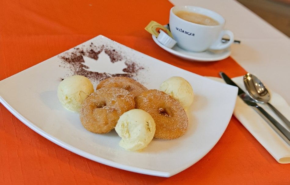 Three donuts with vanilla ice cream scoops on a plate, decorated with cocoa powder. In the background, a cup of coffee on a table with an orange tablecloth., © Hotel Neuwirtshaus Three donuts with vanilla ice cream scoops on a plate, decorated with cocoa powder. In the background, a cup of coffee on a table with an orange tablecloth., © Hotel Neuwirtshaus