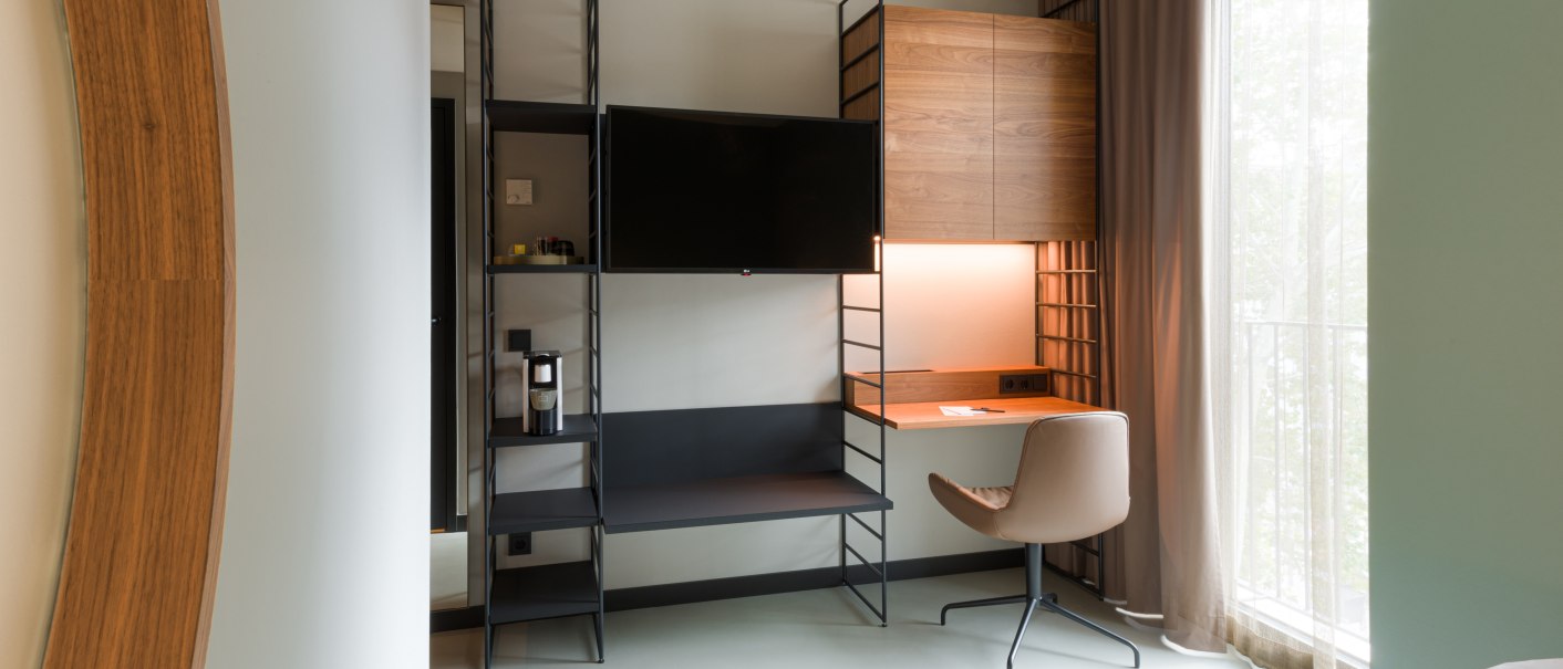 Modernes Zimmer mit Schreibtisch, Stuhl, Fernseher und großem Fenster. Helle, minimalistische Einrichtung mit Holz- und Metallelementen., © Emilu Hotel GmbH