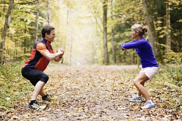 Zwei Personen trainieren im Wald, machen Kniebeugen auf einem mit Laub bedeckten Weg. Sie tragen sportliche Kleidung und lächeln sich an., © Waldhotel Stuttgart GmbH Zwei Personen trainieren im Wald, machen Kniebeugen auf einem mit Laub bedeckten Weg. Sie tragen sportliche Kleidung und lächeln sich an., © Waldhotel Stuttgart GmbH