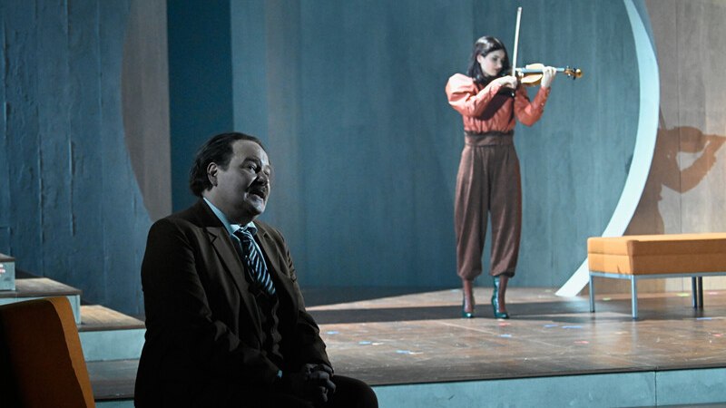 Ein Mann im Anzug sitzt auf einer Bühne, während eine Frau im Hintergrund Geige spielt. Die Szene wirkt dramatisch und konzentriert., © Württembergische Staatstheater Stuttgart Ein Mann im Anzug sitzt auf einer Bühne, während eine Frau im Hintergrund Geige spielt. Die Szene wirkt dramatisch und konzentriert., © Württembergische Staatstheater Stuttgart