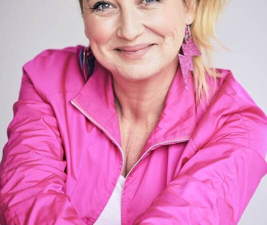 A woman with blonde hair wears a pink jacket and smiles friendly into the camera. She has eye-catching earrings and bracelets., &copy; Rosenau Kultur e.V.