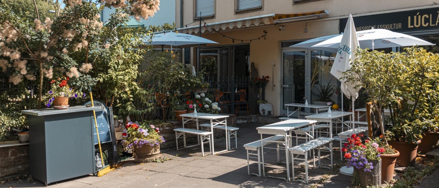 Cosy outdoor area of a café with white tables and chairs, surrounded by lush plants and colorful flowers., © SMG Stuttgart Marketing GmbH - Sarah Schmid