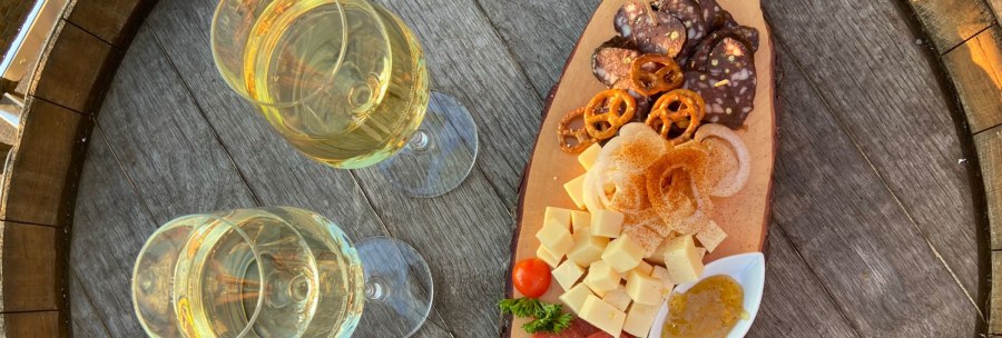 Zwei Gläser Weißwein neben einer Holzplatte mit Käsewürfeln, Wurst, Brezeln, Zwiebeln und Dip auf einem rustikalen Holztisch., © Weinhof Zaiß