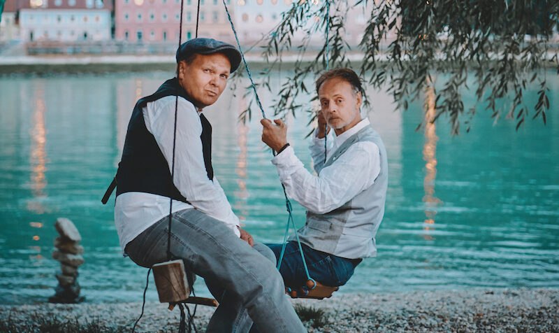 Two men are sitting on swings on the riverbank, elegantly dressed. Water and buildings can be seen in the background., &copy; Rosenau Kultur e.V.