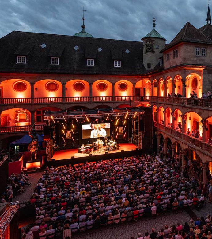 Open-Air-Konzert im Innenhof eines historischen Gebäudes, beleuchtet in Rot. Zuschauer sitzen auf Balkonen und im Innenhof, Bühne mit Musikern., © jazzopen stuttgart, Reiner Pfisterer