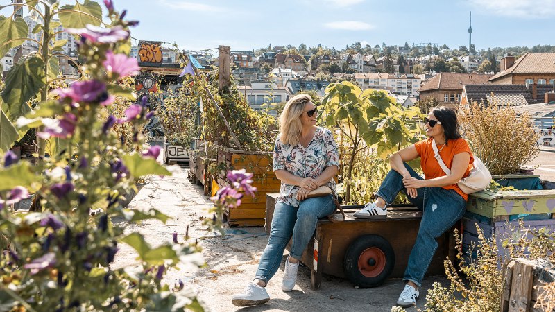 Zwei Frauen sitzen in einem urbanen Dachgarten mit Pflanzen und genießen den Ausblick auf die Stadt im Hintergrund., © © Stuttgart-Marketing GmbH, Sarah Schmid Zwei Frauen sitzen in einem urbanen Dachgarten mit Pflanzen und genießen den Ausblick auf die Stadt im Hintergrund., © © Stuttgart-Marketing GmbH, Sarah Schmid
