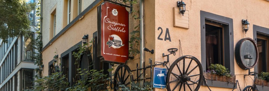 Building with the sign 'Stuttgarter Stäffele', decorated with wagon wheels and plants. The façade is beige with black window frames., © SMG Stuttgart Marketing GmbH - Sarah Schmid Building with the sign 'Stuttgarter Stäffele', decorated with wagon wheels and plants. The façade is beige with black window frames., © SMG Stuttgart Marketing GmbH - Sarah Schmid