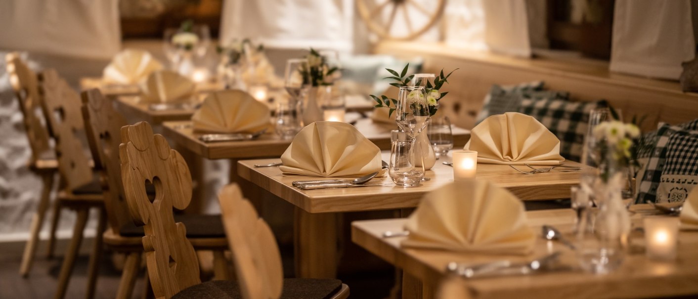 Ein gemütliches Restaurant mit Holzmöbeln, elegant gedeckten Tischen, gefalteten Servietten und Kerzenlicht. Im Hintergrund ein Wagenrad an der Wand., © TOMAS