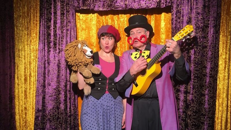 Zwei verkleidete Personen stehen vor einem bunten Vorhang. Die Frau h&auml;lt eine L&ouml;wenpuppe, der Mann spielt Gitarre und tr&auml;gt einen Zylinder., &copy; Theaterhaus Stuttgart e.V.