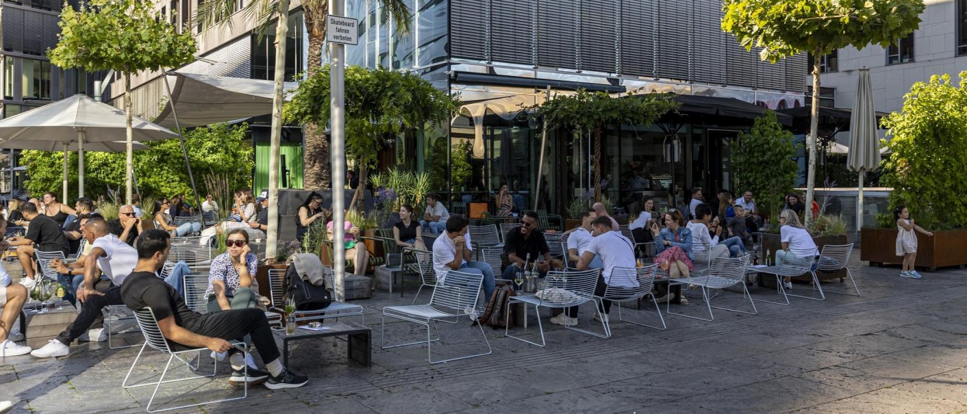 Menschen entspannen sich in einem belebten Café im Freien, umgeben von Pflanzen und modernen Gebäuden. Ein Schild verbietet Skateboardfahren., © SMG, Sarah Schmid