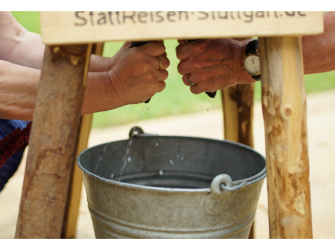Zwei Personen melken eine Attrappe über einem Eimer bei einem Outdoor-Event., © Cool-Tours StattReisen Zwei Personen melken eine Attrappe über einem Eimer bei einem Outdoor-Event., © Cool-Tours StattReisen