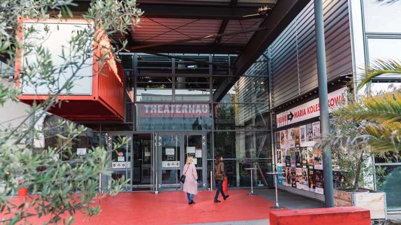 Entrance to the Theaterhaus Stuttgart with red floor, glass facade and posters. Two people enter the building. Plants and modern architecture are visible., © Stuttgart-Marketing GmbH, wpsteinheisser