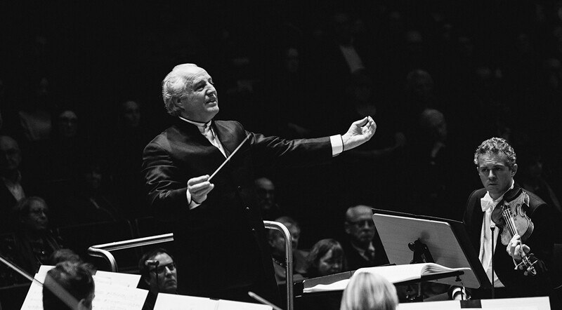 Ein Dirigent führt ein Orchester mit erhobenem Taktstock. Ein Geiger sitzt rechts, das Publikum ist im Hintergrund sichtbar., © Württembergische Staatstheater Stuttgart Ein Dirigent führt ein Orchester mit erhobenem Taktstock. Ein Geiger sitzt rechts, das Publikum ist im Hintergrund sichtbar., © Württembergische Staatstheater Stuttgart