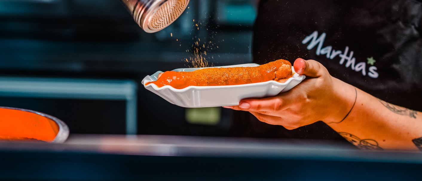Eine Currywurst wird mit Gewürz bestreut. Die Person trägt eine Schürze mit der Aufschrift "Martha's"., © Emma von Bergenspitz Eine Currywurst wird mit Gewürz bestreut. Die Person trägt eine Schürze mit der Aufschrift "Martha's"., © Emma von Bergenspitz