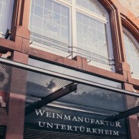 Fassade der Weinmanufaktur Untertürkheim mit Glasvordach, Schriftzug und dekorativen Fenstern., © Weinmanufaktur Stuttgart