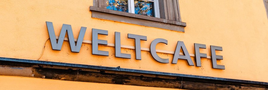 The words 'Weltcafé' are emblazoned in large letters on a yellow wall, below a window with a blue frame., © SMG Stuttgart Marketing GmbH - Sarah Schmid