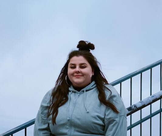 A woman in a blue hooded jacket stands smiling on a staircase. The sky is cloudy and the railing is visible., &copy; Rosenau Kultur e.V.