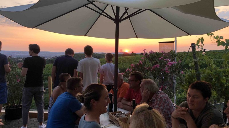 Menschen sitzen unter Sonnenschirmen in einem Weinberg und genießen den Sonnenuntergang. Einige stehen und blicken in die Ferne., © Weingut Diehl