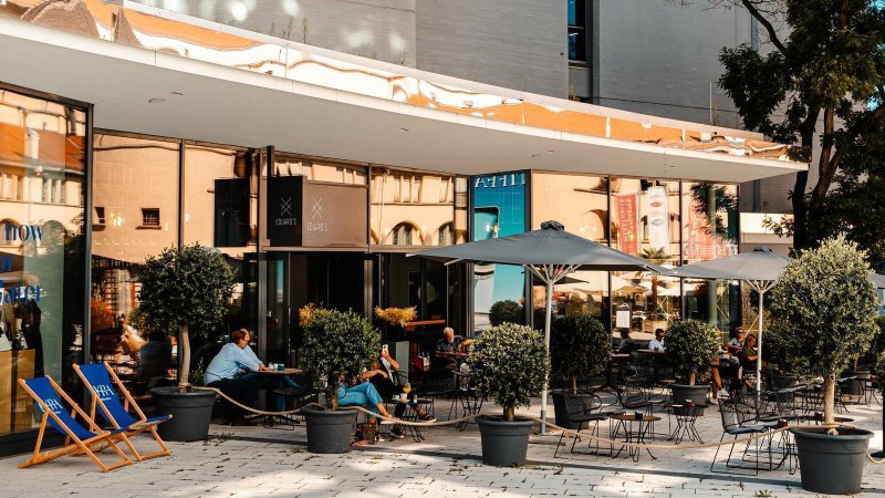 Außenbereich eines Cafés mit Tischen, Stühlen und Sonnenschirmen. Menschen sitzen und entspannen. Pflanzen in Töpfen dekorieren den Bereich., © Stuttgart Marketing GmbH, Sarah Schmid Außenbereich eines Cafés mit Tischen, Stühlen und Sonnenschirmen. Menschen sitzen und entspannen. Pflanzen in Töpfen dekorieren den Bereich., © Stuttgart Marketing GmbH, Sarah Schmid