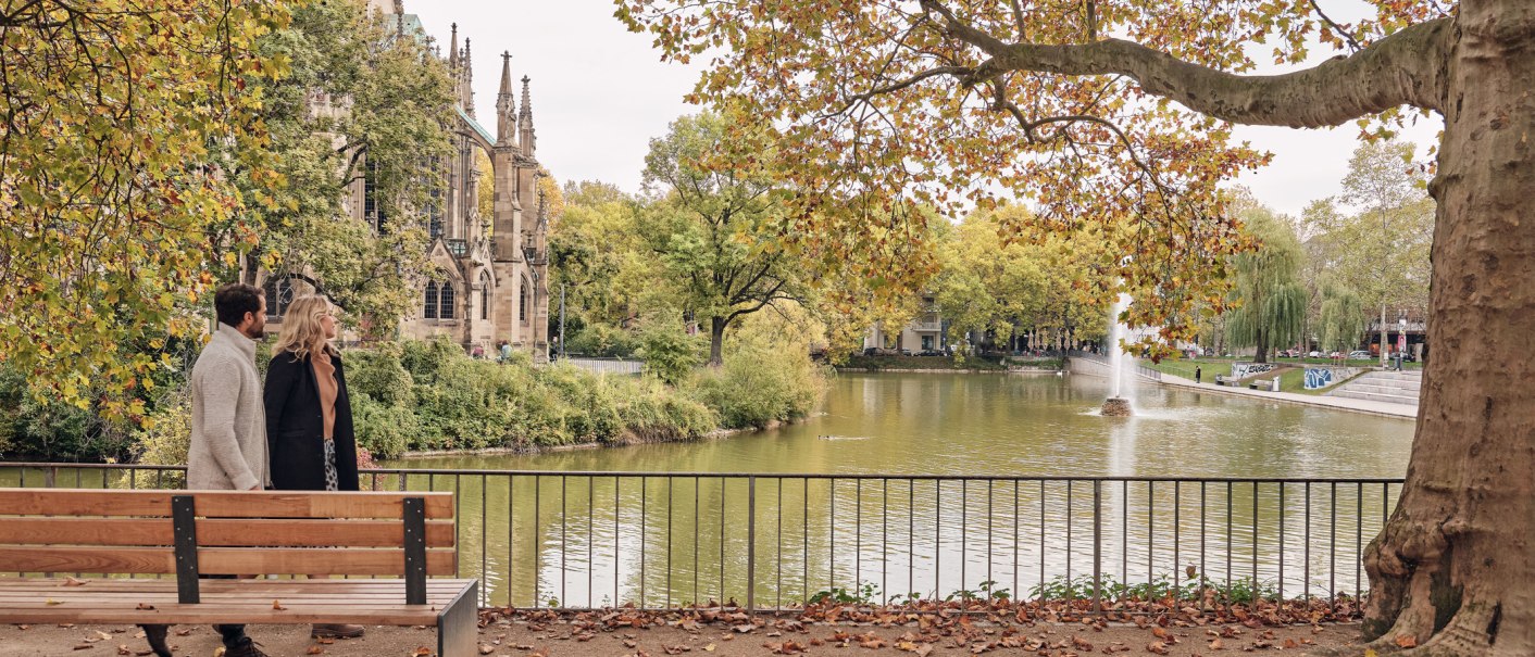 Ein Paar spaziert am Feuersee entlang, im Hintergrund die Johanneskirche und herbstliche Bäume. Ein Brunnen sprudelt im See., © SMG Christoph Düpper