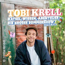 Ein Mann sitzt l&auml;chelnd vor einer Holzh&uuml;tte, umgeben von B&auml;umen. Oben steht der Titel der Show: 'Tobi Krell - R&auml;tsel, Wissen, Abenteuer - Die gro&szlig;e Sommershow'., &copy; links im Bild