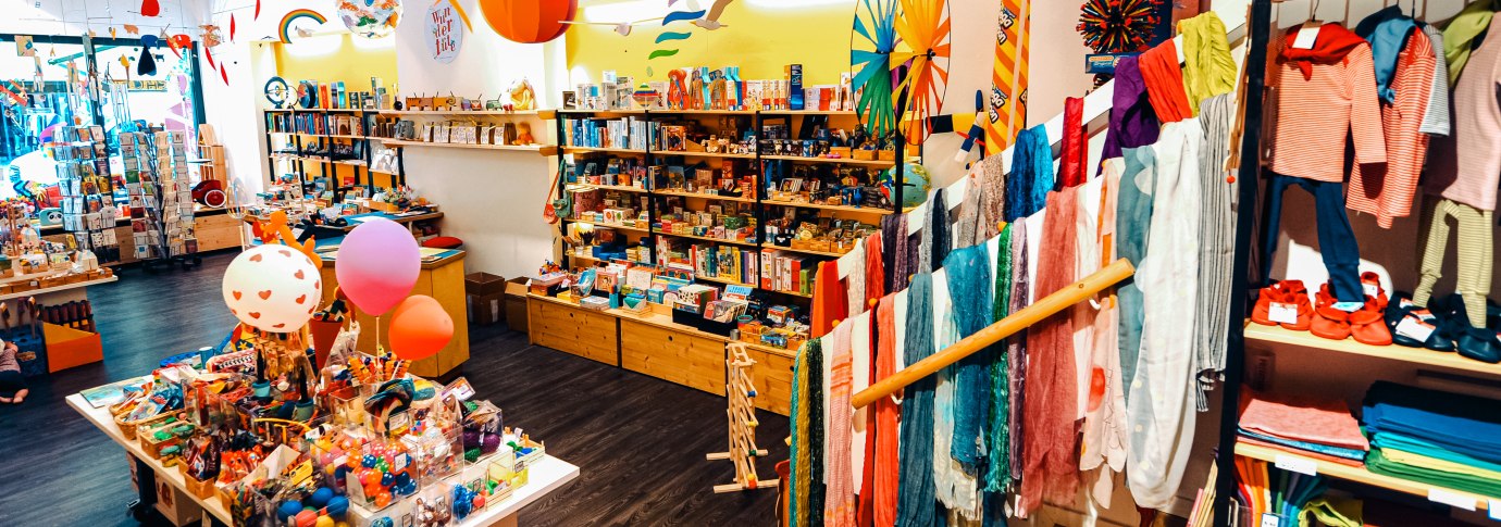 Ein bunter Spielzeugladen mit Regalen voller Spielsachen, Kleidung und Dekorationen. Bunte Schals und Spielzeuge sind prominent ausgestellt., &copy; 1000sch&ouml;n