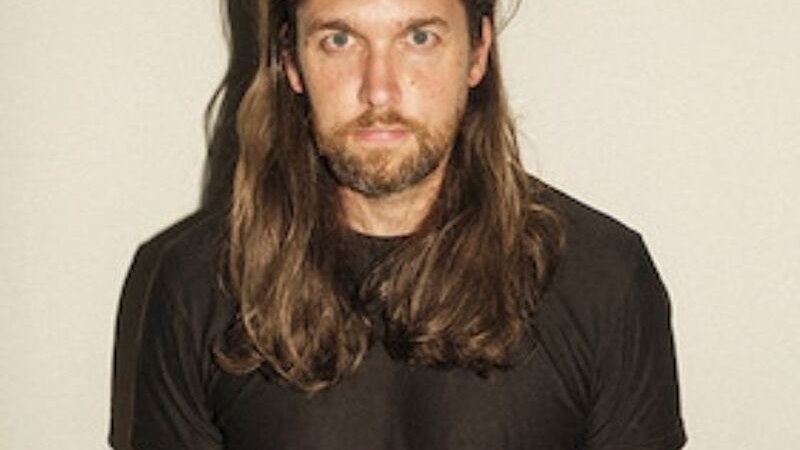 Ein Mann mit langen Haaren und Bart trägt ein schwarzes T-Shirt und steht vor einem neutralen Hintergrund., © Theaterhaus Stuttgart e.V. Ein Mann mit langen Haaren und Bart trägt ein schwarzes T-Shirt und steht vor einem neutralen Hintergrund., © Theaterhaus Stuttgart e.V.