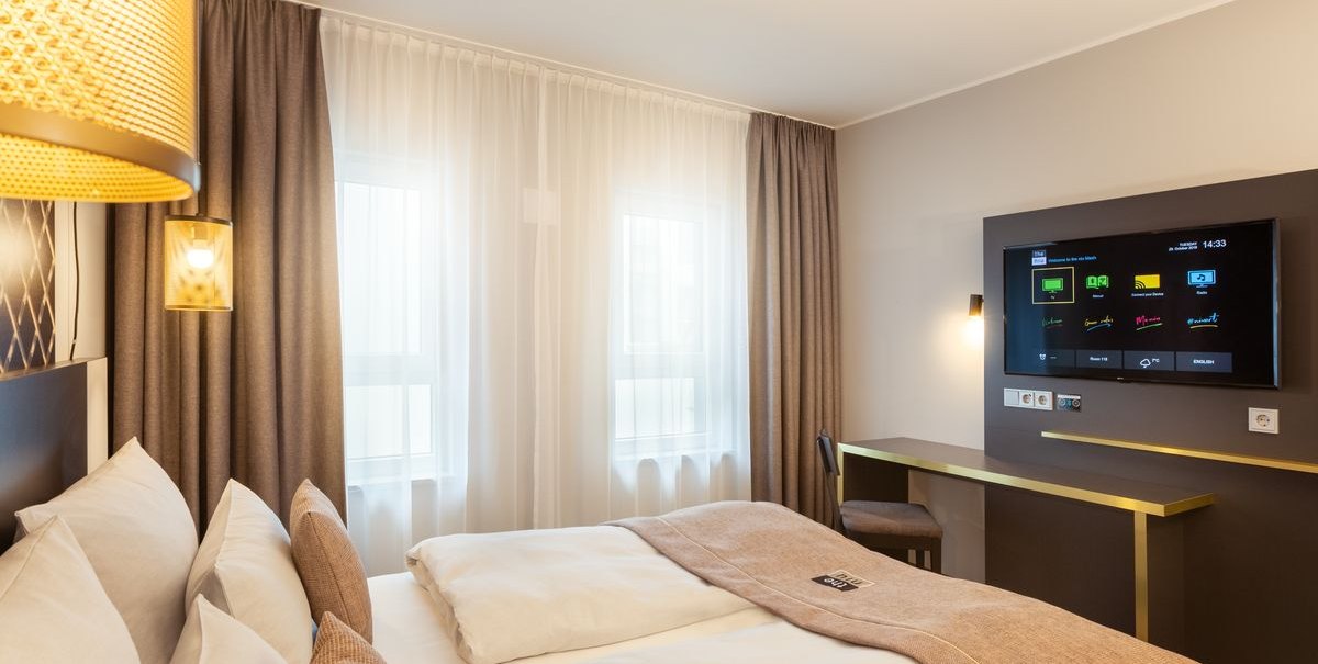 Modernes Hotelzimmer mit Doppelbett, TV an der Wand und kleinem Schreibtisch. Große Fenster mit Vorhängen lassen Tageslicht herein., © Novum Hospitality