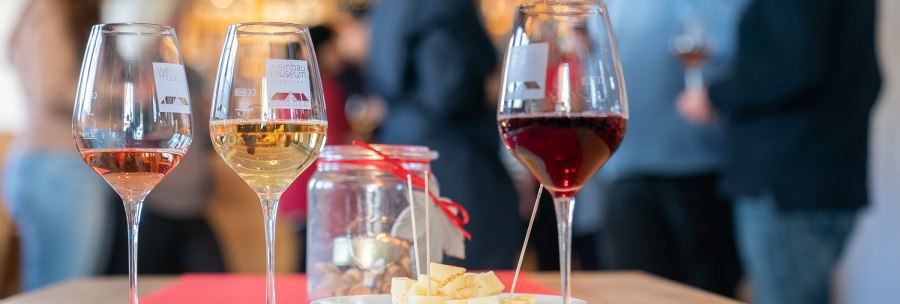 Three wine glasses with red, white and rosé wine stand on a table. Next to them is a plate with cheese cubes and a glass with a candle. People in the background., © Thomas Niedermüller Three wine glasses with red, white and rosé wine stand on a table. Next to them is a plate with cheese cubes and a glass with a candle. People in the background., © Thomas Niedermüller